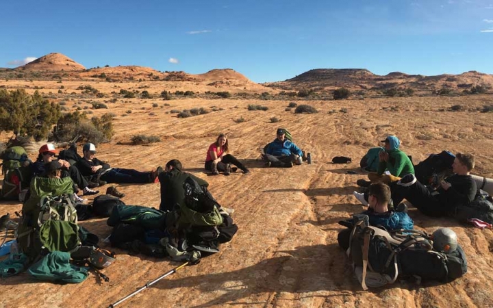 A group of people take a break from backpacking and sit in a desert landscape.